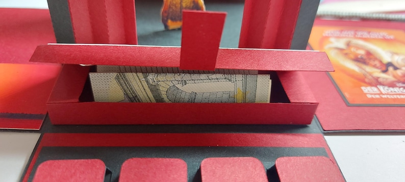 Puede incluir: Una manualidad de papel rojo y negro con una peque&ntilde;a caja rectangular que contiene un billete enrollado. La caja forma parte de una estructura m&aacute;s grande y abierta con detalles rojos y negros. Un anuncio de comida es visible en el fondo.