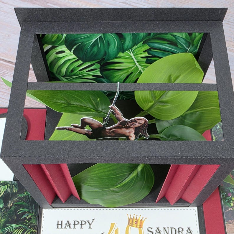 Puede incluir: Una tarjeta negra hecha a mano con un tema de jungla. La tarjeta presenta un recorte de una persona balance&aacute;ndose en una liana, rodeada de hojas verdes y un fondo tropical. La tarjeta tambi&eacute;n incluye el texto "HAPPY 40 SANDRA".