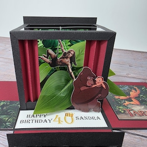 Puede incluir: Una tarjeta de cumplea&ntilde;os emergente hecha a mano con un tema de jungla. La tarjeta presenta im&aacute;genes de Tarz&aacute;n, un gorila sosteniendo a un beb&eacute; y follaje tropical. La tarjeta dice "Happy Birthday 40 Sandra". La tarjeta es negra, roja y verde.
