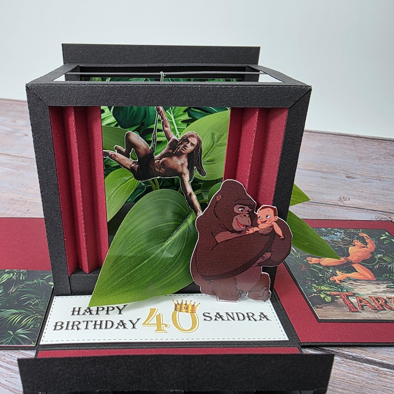 Puede incluir: Una tarjeta de cumplea&ntilde;os emergente hecha a mano con un tema de jungla. La tarjeta presenta im&aacute;genes de Tarz&aacute;n, un gorila sosteniendo a un beb&eacute; y follaje tropical. La tarjeta dice "Happy Birthday 40 Sandra". La tarjeta es negra, roja y verde.