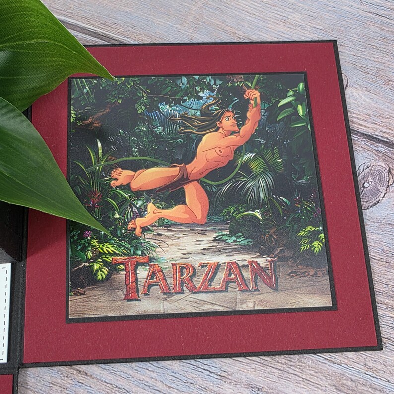 Puede incluir: Impresi&oacute;n enmarcada de Tarz&aacute;n balance&aacute;ndose por una jungla. La imagen tiene un borde rojo y la palabra "TARZAN" en rojo en la parte inferior. El fondo es una exuberante jungla verde.