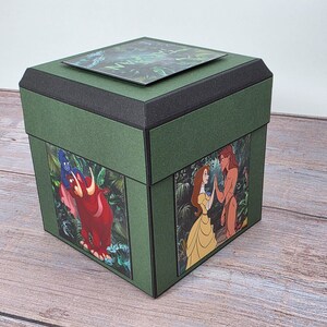 Puede incluir: Una caja decorativa verde con ribetes negros y una tapa. La caja presenta im&aacute;genes de personajes animados en los laterales y la palabra "TARZAN" en la tapa. La caja es de forma cuadrada.