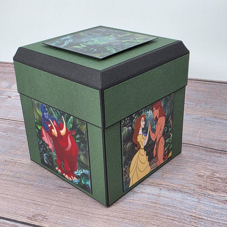 Puede incluir: Una caja decorativa verde con ribetes negros y una tapa. La caja presenta im&aacute;genes de personajes animados en los laterales y la palabra "TARZAN" en la tapa. La caja es de forma cuadrada.