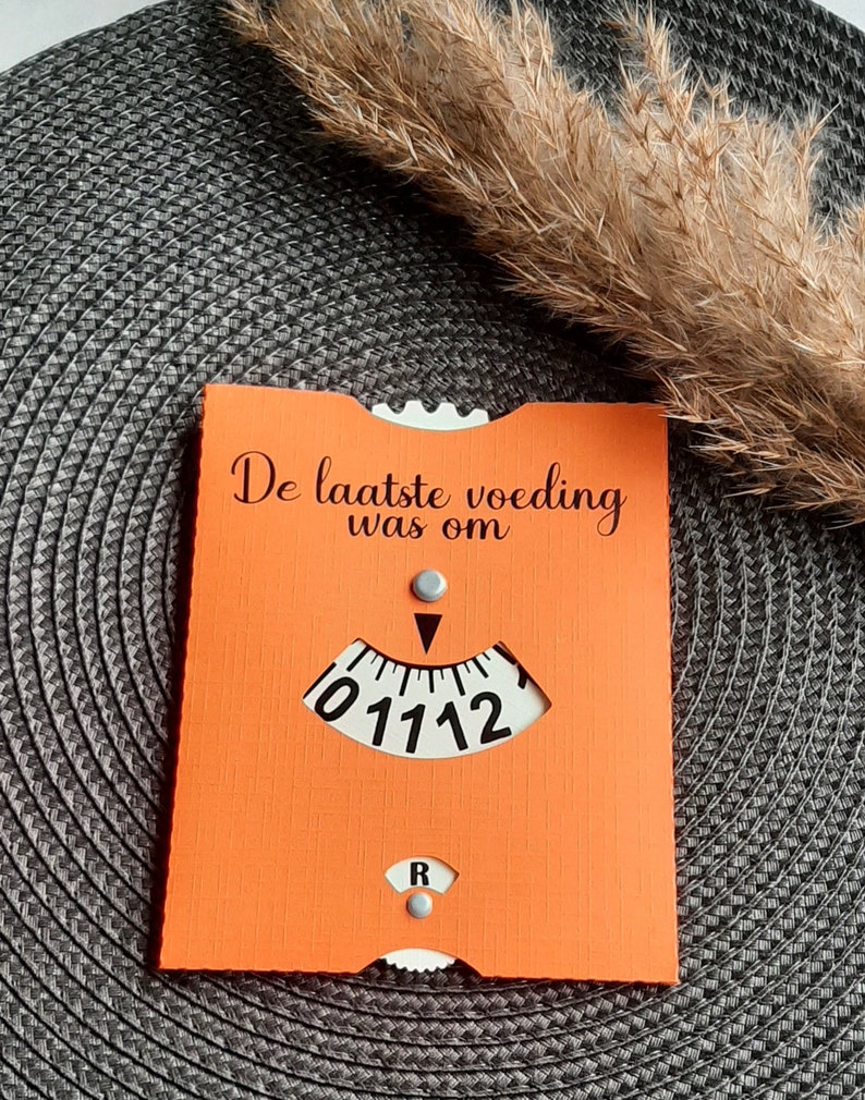 May include: Orange card with the text "De laatste voeding was om" and a white dial with black numbers and a black arrow. The dial reads "01112".