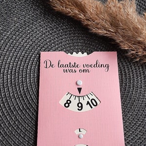 May include: A pink card with the text "De laatste voeding was om" and a dial with numbers 8, 9, and 10. The dial has a white arrow pointing to the number 9.
