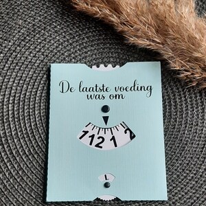 May include: A light blue card with the text "De laatste voeding was om" and a black and white dial with numbers 1, 2, 12, and 7.