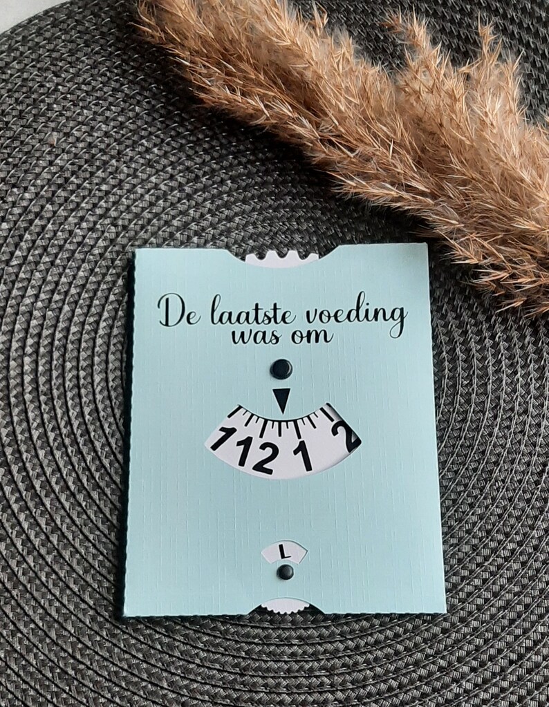 May include: A light blue card with the text "De laatste voeding was om" and a black and white dial with numbers 1, 2, 12, and 7.