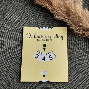 May include: A yellow card with the text "De laatste voeding was om" and a black and white dial with numbers 3, 4, and 5. The dial has a black arrow pointing to the number 4.