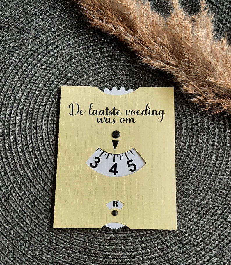 May include: A yellow card with the text "De laatste voeding was om" and a black and white dial with numbers 3, 4, and 5. The dial has a black arrow pointing to the number 4.