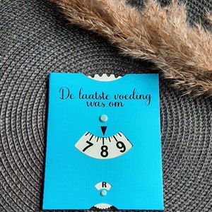 May include: A blue card with the text "De laatste voeding was om" and a white dial with black numbers 7, 8, and 9. The dial has a black arrow pointing to the number 8.