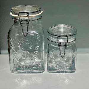 May include: Two clear glass jars with metal hinged lids. The jars have a square shape and embossed wheat sheaf design. The larger jar has a white rubber seal. The jars are of different sizes, suitable for kitchen storage.