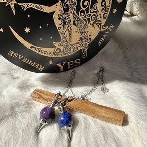 May include: Two pendulum dowsing charms with a silver chain. The charms are made of amethyst and lapis lazuli. The charms are hanging from a wooden stick. The background is a white fuzzy surface. The charms are used for divination.