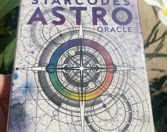 May include: A 56-card deck and guidebook for the Starcodes Astro Oracle. The box features a colorful, geometric design with a rainbow circle and a star in the center. The text "Starcodes Astro Oracle" is printed in purple on the front of the box.