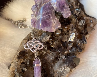 Puede incluir: Figurita de unicornio de amatista con una cadena de plata y un colgante con un nudo en forma de corazón y una punta de amatista. El unicornio y el colgante se muestran sobre un lecho de cristales de cuarzo negro y transparente.