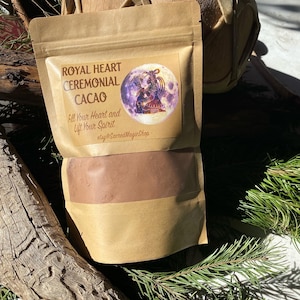 May include: A brown paper bag with a label that reads "Royal Heart Ceremonial Cacao" and "Fill Your Heart and Lift Your Spirit" with an image of a woman in a moon. The bag is filled with a brown powder. The bag is sitting on a wooden surface with green pine branches.