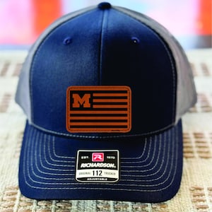 Pode incluir: Boné de caminhoneiro azul marinho com parte traseira em malha cinza. A frente apresenta um patch de couro marrom com um "M" laranja sobre listras laranja e pretas. O boné tem uma aba curva e uma alça ajustável. A etiqueta diz "RICHARDSON 112 TRUCKER".