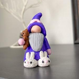 May include: Purple gnome figurine with a gray beard, wearing a pink and purple hat and bunny slippers. The gnome is holding a brown teddy bear.