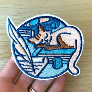 Könnte beinhalten: Gestickter Aufnäher mit einer schlafenden Katze in Blau- und Brauntönen. Die Katze liegt zusammengerollt auf einer blauen Oberfläche, mit einer Feder und anderen Objekten im Hintergrund. Der Aufnäher hat einen weißen Rand.