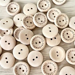 Op de afbeelding: Een verzameling lichtgekleurde houten knopen. Elke knoop heeft twee gaatjes en het woord "handmade" rond een hartsymbool gedrukt. De knopen zijn rond en lijken van natuurlijk hout te zijn.