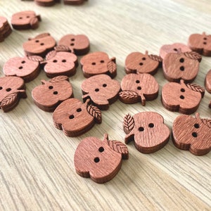 May include: A collection of wooden apple-shaped buttons with two holes each. The buttons are a reddish-brown colour and have a carved leaf detail.