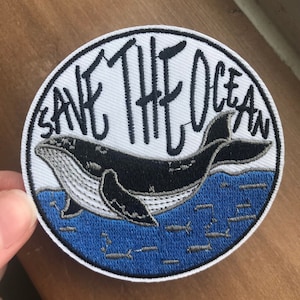 May include: A white, round embroidered patch with a black whale swimming in blue water. The text "Save The Ocean" is written in black around the whale.