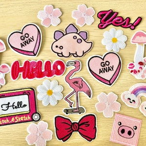 Può includere: Una collezione di toppe termoadesive rosa e bianche con vari disegni, tra cui un fenicottero, un arcobaleno, un fungo, un cuore con il testo "Go Away", un fiore e il testo "Hello".