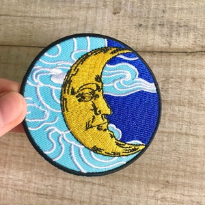 Peut inclure: Une lune croissante jaune avec un visage brodé sur un fond bleu et bleu clair avec des nuages blancs.