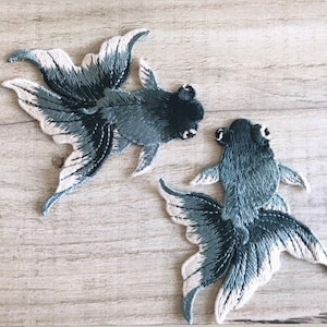 Może przedstawiać: Dwa haftowane naszywki do prasowania z czarnymi i niebieskimi złotymi rybkami z białymi płetwami. Naszywki znajdują się na jasnym drewnianym tle.