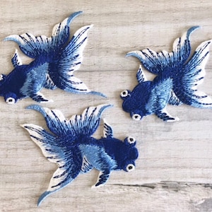 May include: Three embroidered goldfish patches. The fish are mainly navy blue with white and light blue detailing in their fins and tails. They are displayed on a light wood-grain surface.