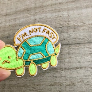 Può includere: Una toppa ricamata bianca con una tartaruga verde e il testo "Non sono veloce".