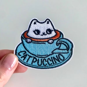 May include: A blue and white embroidered patch featuring a white cat sitting in a blue coffee cup. The text "Cat-puccino" is embroidered in black around the cup.