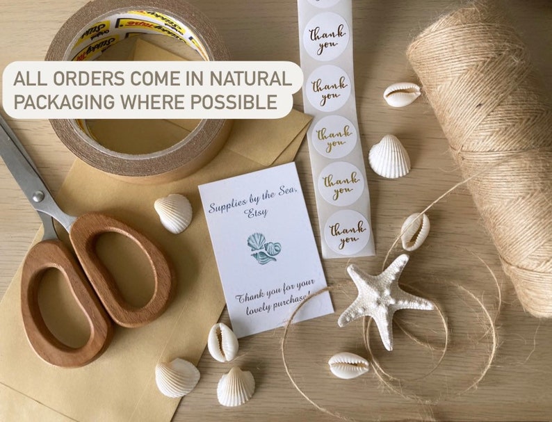 May include: An overhead shot of packaging materials and decorative elements. Includes scissors, tape, twine, seashells, stickers with "thank you" text, and a card that reads "Supplies by the Sea, Etsy". The text "ALL ORDERS COME IN NATURAL PACKAGING WHERE POSSIBLE" is also visible.