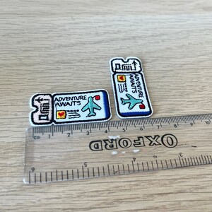 May include: Two embroidered patches in the shape of airplane tickets. The patches are white with blue, red, and yellow accents. The text "ADVENTURE AWAITS" is visible on both patches, along with an airplane graphic. The patches are approximately 1.5 inches and 2.5 inches in length.