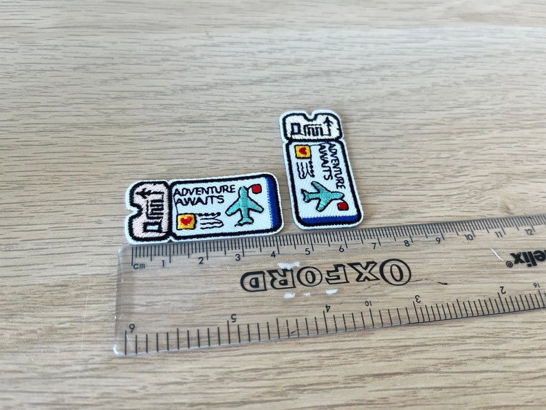 May include: Two embroidered patches in the shape of airplane tickets. The patches are white with blue, red, and yellow accents. The text "ADVENTURE AWAITS" is visible on both patches, along with an airplane graphic. The patches are approximately 1.5 inches and 2.5 inches in length.