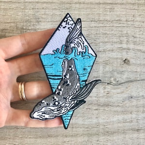以下が含まれることがあります： 水から飛び出すクジラの刺繍パッチ。ダイヤモンド型で、青と白の背景。クジラは青とグレーです。