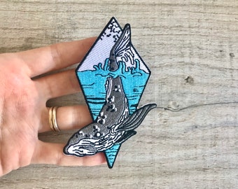 Può includere: Una toppa ricamata con una balena che salta fuori dall'acqua, in forma di diamante con uno sfondo blu e bianco. La balena è blu e grigia.