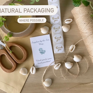May include: A close-up of a brown paper envelope with a pair of wooden handled scissors, a roll of brown twine, and a small card that reads "Supplies by the Sea, Etsy" and "Thank you for your lovely purchase!"  The card is decorated with a small illustration of seashells.  There are also several small white seashells and a sheet of round stickers that say "Thank you" in gold lettering.