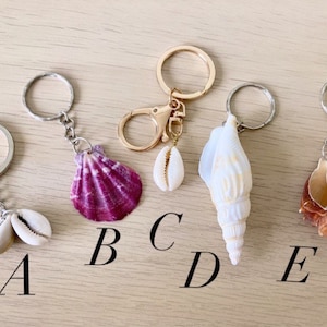 Puede incluir: Cinco llaveros diferentes con diferentes conchas marinas adjuntas. El llavero A presenta dos conchas de cauri blancas. El llavero B tiene una concha púrpura. El llavero C tiene una concha de cauri blanca. El llavero D tiene una concha blanca. El llavero E tiene una concha marrón.