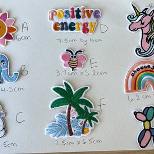 May include: A collection of embroidered patches featuring various designs. Includes a smiling sun, a colorful elephant, a cat with a floral crown, the words "Positive Energy", a butterfly, palm trees, a unicorn, a rainbow with "choose joy", and a balloon dog.
