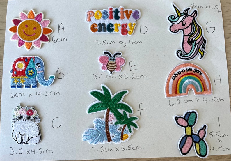 May include: A collection of embroidered patches featuring various designs. Includes a smiling sun, a colorful elephant, a cat with a floral crown, the words "Positive Energy", a butterfly, palm trees, a unicorn, a rainbow with "choose joy", and a balloon dog.