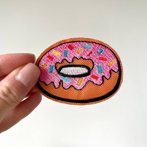 Può includere: Toppa ricamata di una ciambella con glassa rosa e zuccherini colorati. La ciambella ha un contorno marrone e un centro bianco. La toppa è su sfondo bianco.