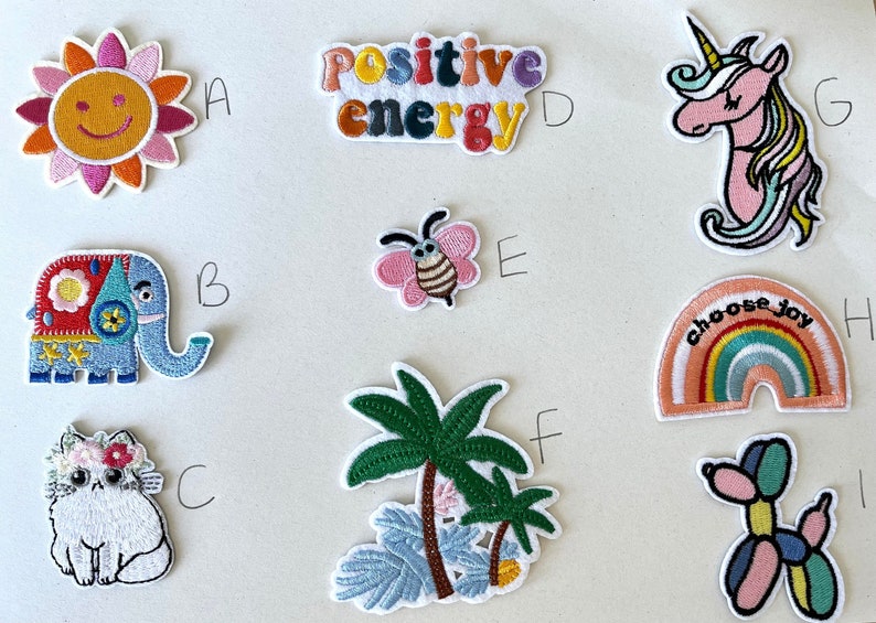 May include: Collection of embroidered patches featuring various designs. Includes a smiling sun, an elephant, a cat with flowers, the words "Positive Energy", a butterfly, palm trees, a unicorn, a rainbow with "choose joy", and a balloon dog.