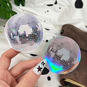 Könnte beinhalten: Zwei kleine, runde Discokugeln mit verspiegelten Oberflächen. Eine Kugel wird in einer Hand mit einem Stern-Nageldesign gehalten. Die Discokugeln reflektieren das Licht und erzeugen einen schimmernden Effekt. Der Hintergrund ist ein weißer Stoff mit schwarzen Tupfen.