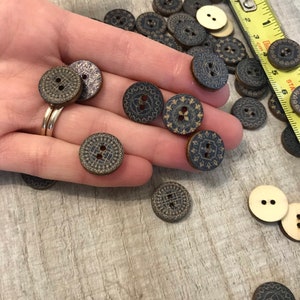 May include: A hand holding a variety of wooden buttons with a black and gold geometric design. The buttons are approximately 1 inch in diameter and have two holes.