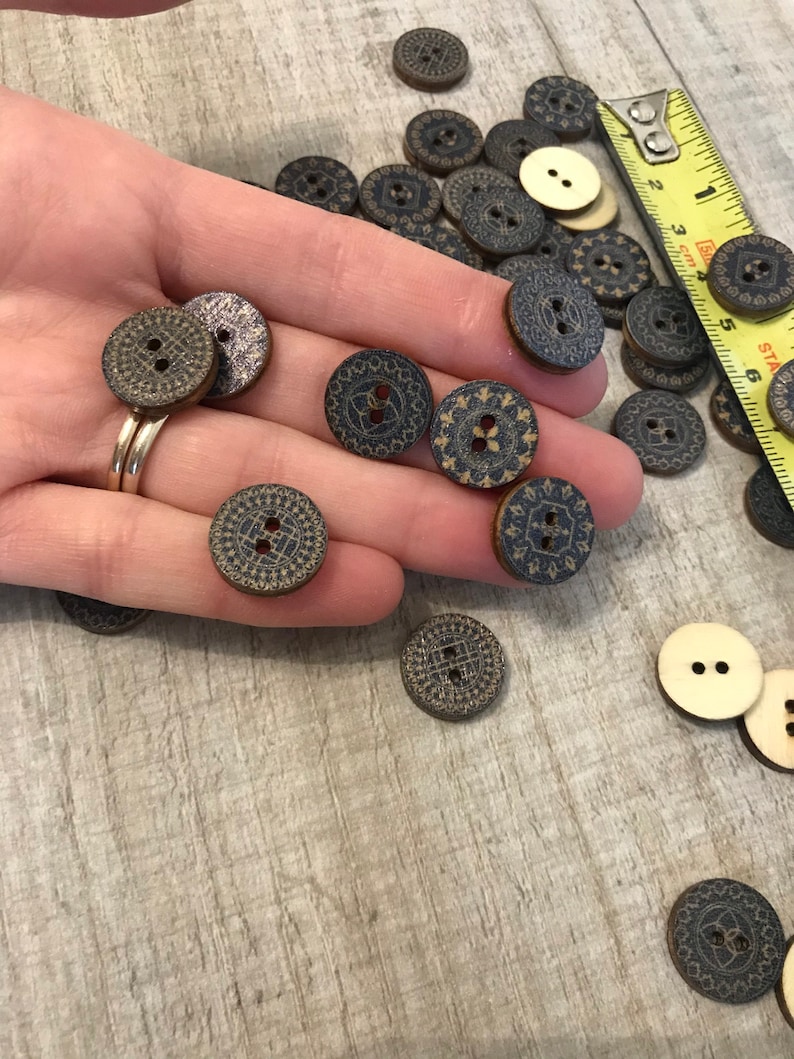 May include: A hand holding a variety of wooden buttons with a black and gold geometric design. The buttons are approximately 1 inch in diameter and have two holes.
