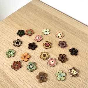 Puede incluir: Una colección de botones de madera con forma de flor en varios colores, incluyendo verde, rosa, marrón y naranja. Los botones están dispuestos sobre una superficie de madera clara, mostrando sus intrincados diseños florales y texturas. Ideal para proyectos de costura y manualidades.