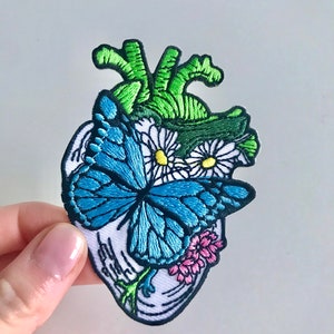 以下が含まれることがあります： 刺繍されたワッペン。解剖学的な心臓のデザインで、青い蝶、白いデイジー、ピンクの花、緑のアクセントが特徴です。ワッペンは黒い縁取りで、白い背景にあります。