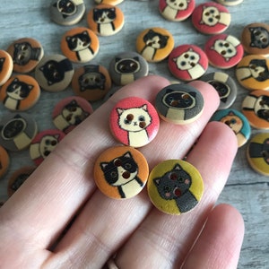 May include: A collection of wooden buttons featuring various cat designs. The buttons are painted in a variety of colors, including orange, yellow, pink, and brown. Some of the cats have black eyes, while others have blue eyes. The buttons are all about 1 inch in diameter.