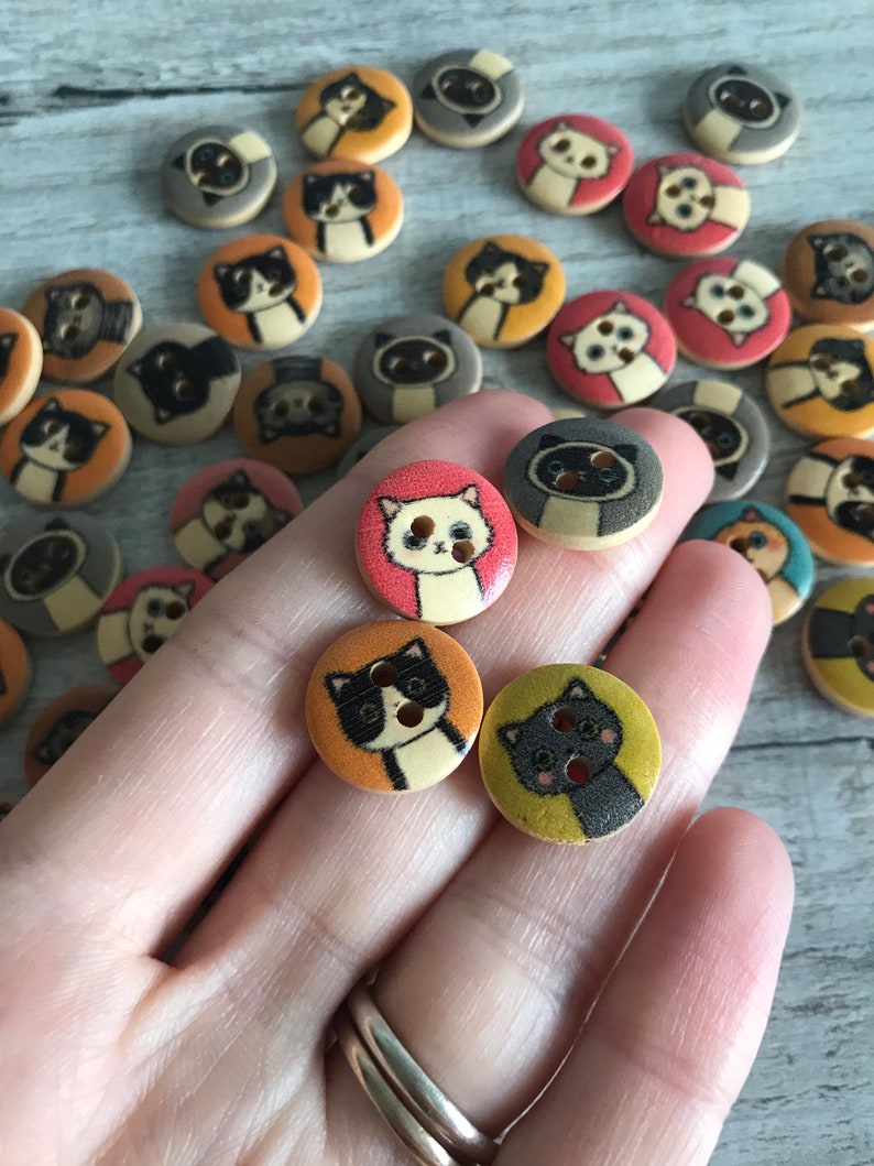May include: A collection of wooden buttons featuring various cat designs. The buttons are painted in a variety of colors, including orange, yellow, pink, and brown. Some of the cats have black eyes, while others have blue eyes. The buttons are all about 1 inch in diameter.