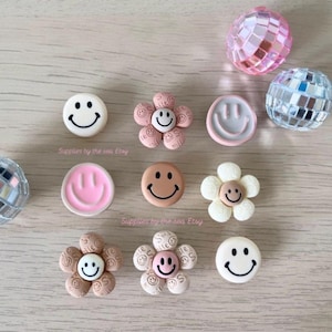 May include: A collection of decorative items featuring smiley face buttons in white, pink, and brown, and flower-shaped buttons with smiley faces. Also, two silver and one pink disco ball ornaments are visible.